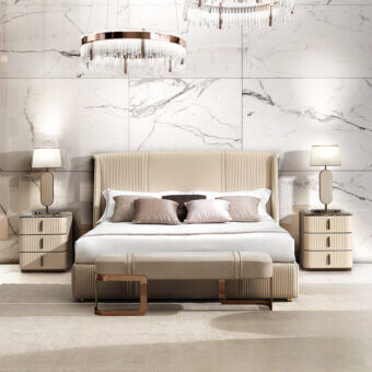 King bed with beige upholstered headboard, rose gold bench, dark grey & beige nightstands, acrylic chandelier, and marble tile backdrop.