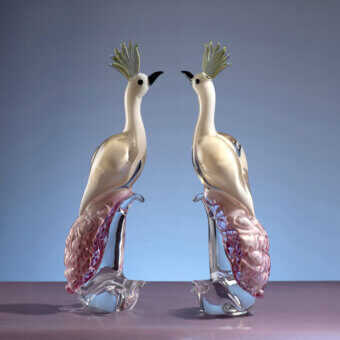 Crystal peacock sculptures, clear glass with pearlescent white bodies, pink & purple filaments, on mauve surface against blue-lavender gradient.